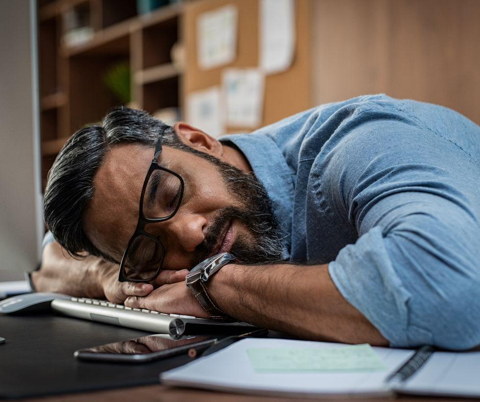 A man wearing glasses and a blue shirt asleep on his desk, leaning his head on his arms over a computer keyboard, illustrating writer's fatigue and the need for a pause.